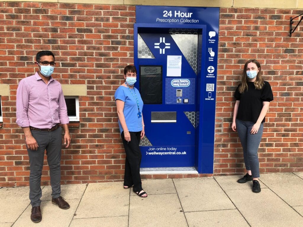 Our pharmacy team gathered around the blue 24/7 prescription collection point. Three staff stand around the collection point.