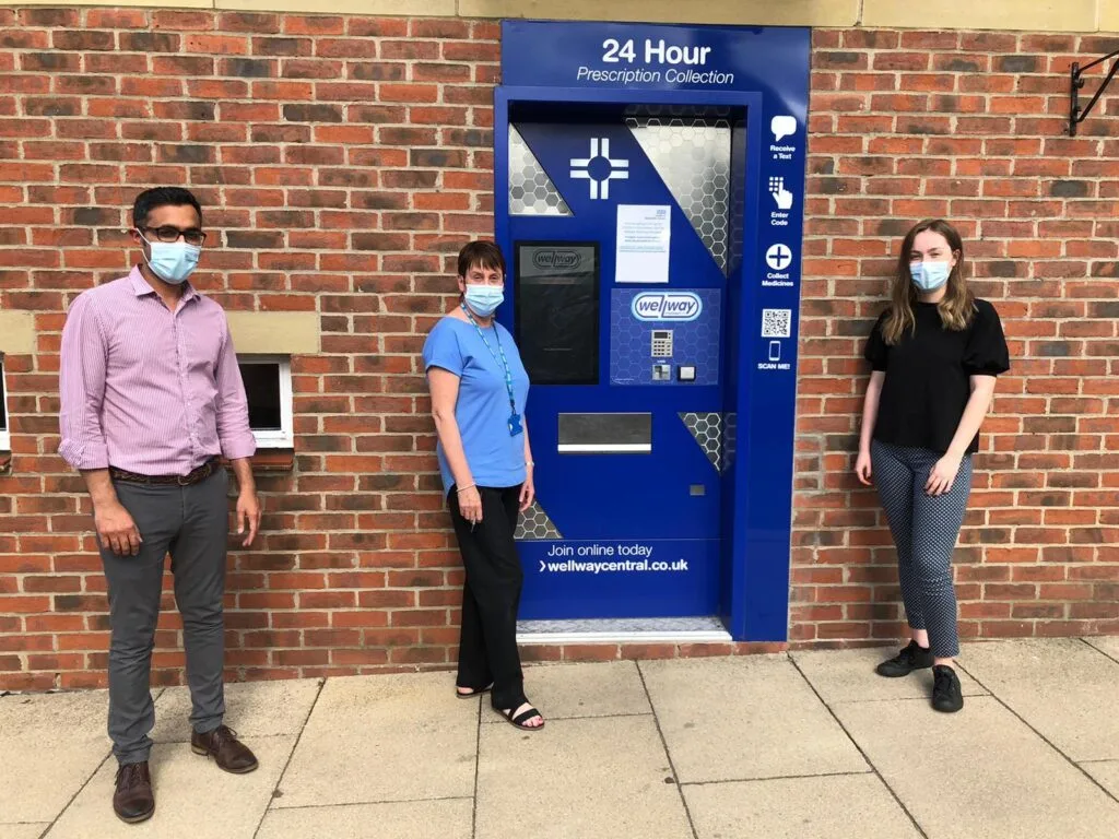 Our pharmacy team gathered around the blue 24/7 prescription collection point. Three staff stand around the collection point.
