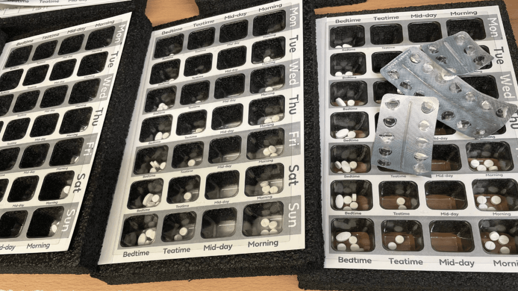 An images of dosette boxes on a counter, open and overlapping each other. Opened blister packs are alongside the boxes.