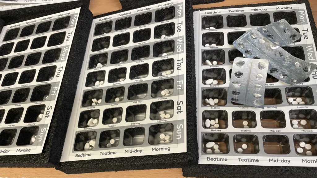 An images of dosette boxes on a counter, open and overlapping each other. Opened blister packs are alongside the boxes.