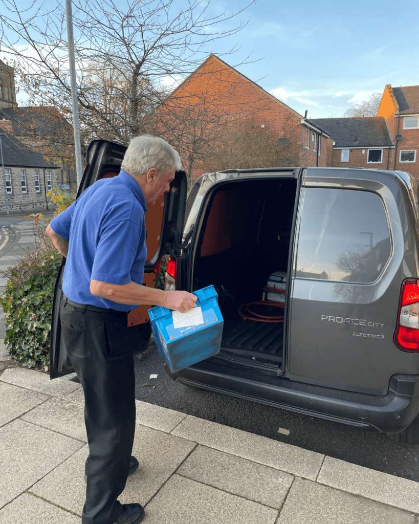 Our delivery driver Duncan loading our grey electric delivery van wearing his blue uniform outside Wellway Morpeth. The rear doors of the van are open as Duncan loads it. Church hall building visible in the background.