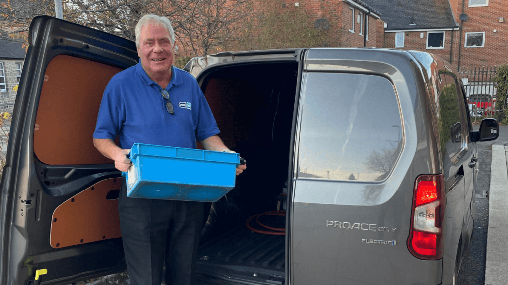 Our prescription delivery driver Duncan beaming in his blue Wellway Uniform, holding a tote box which he is about to load into our electric van.