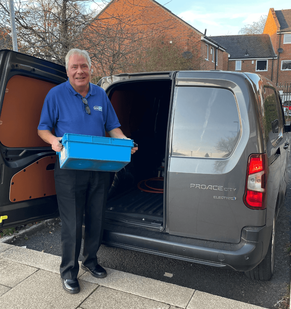 Our prescription delivery driver Duncan beaming in his blue Wellway Uniform, holding a tote box which he is about to load into our electric van.