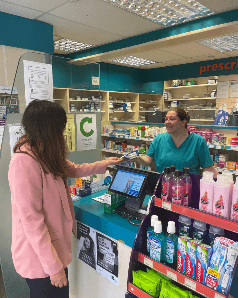 Pharmacy Technician Emily smiling whilst handing out a prescription bag at the pharmacy counter in Ashington Pharmacy. A well-stocked shop is visible in the background.